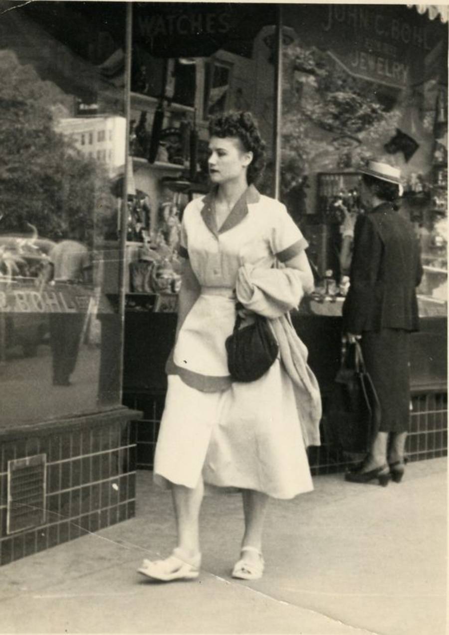 A woman in a light dress walks past a jewelry store window. She carries a coat and purse, while another woman stands by the display. The scene appears to be from the mid-20th century.