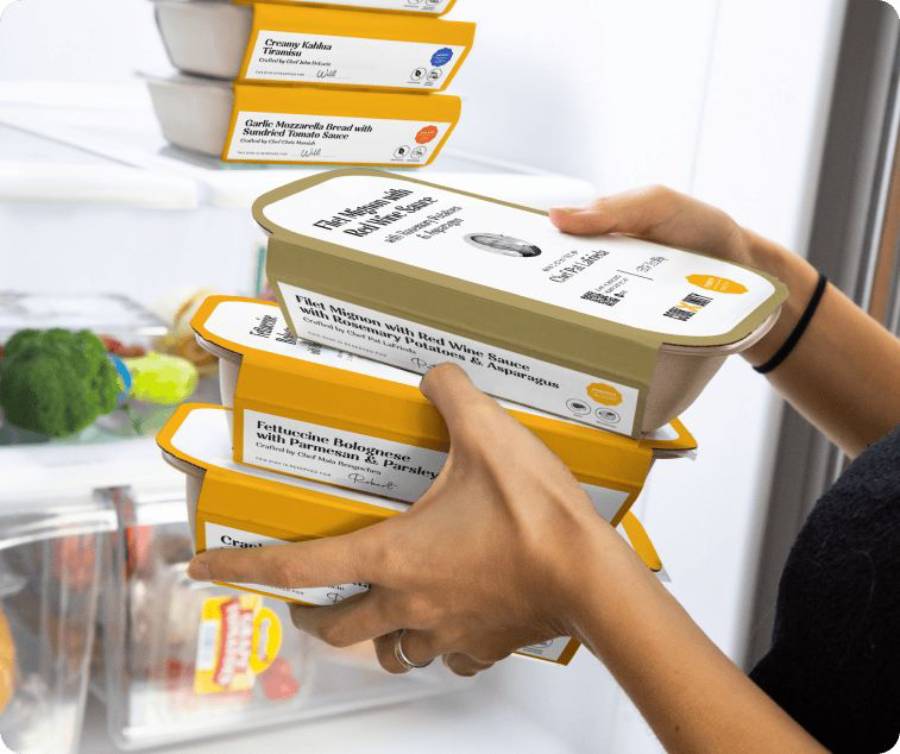 A person places several labeled ready-meal containers into a refrigerator, with visible broccoli and vegetables inside the fridge.