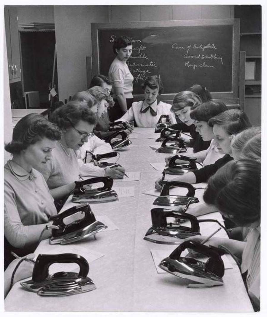 A group of women sit at a long table with irons and notebooks, writing while a teacher stands by a blackboard in a classroom.