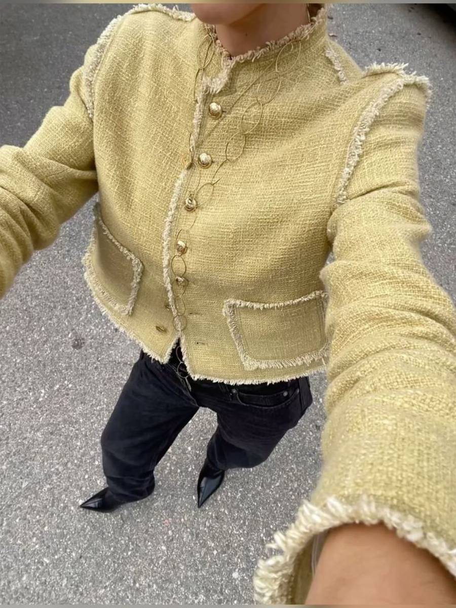 Person wearing a textured light yellow jacket with gold buttons and frayed edges, paired with black pants and pointed black shoes, standing on a gray pavement.