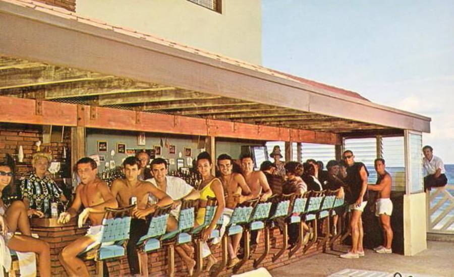A group of people in swimsuits sit and stand around an outdoor bar, enjoying drinks and socializing. The bar is partially covered with a wooden roof and overlooks the ocean on a sunny day.