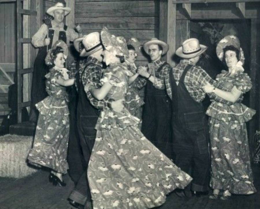 A group of people in matching patterned outfits and straw hats square dancing in pairs inside a rustic wooden building. One man stands in the background watching and smiling.