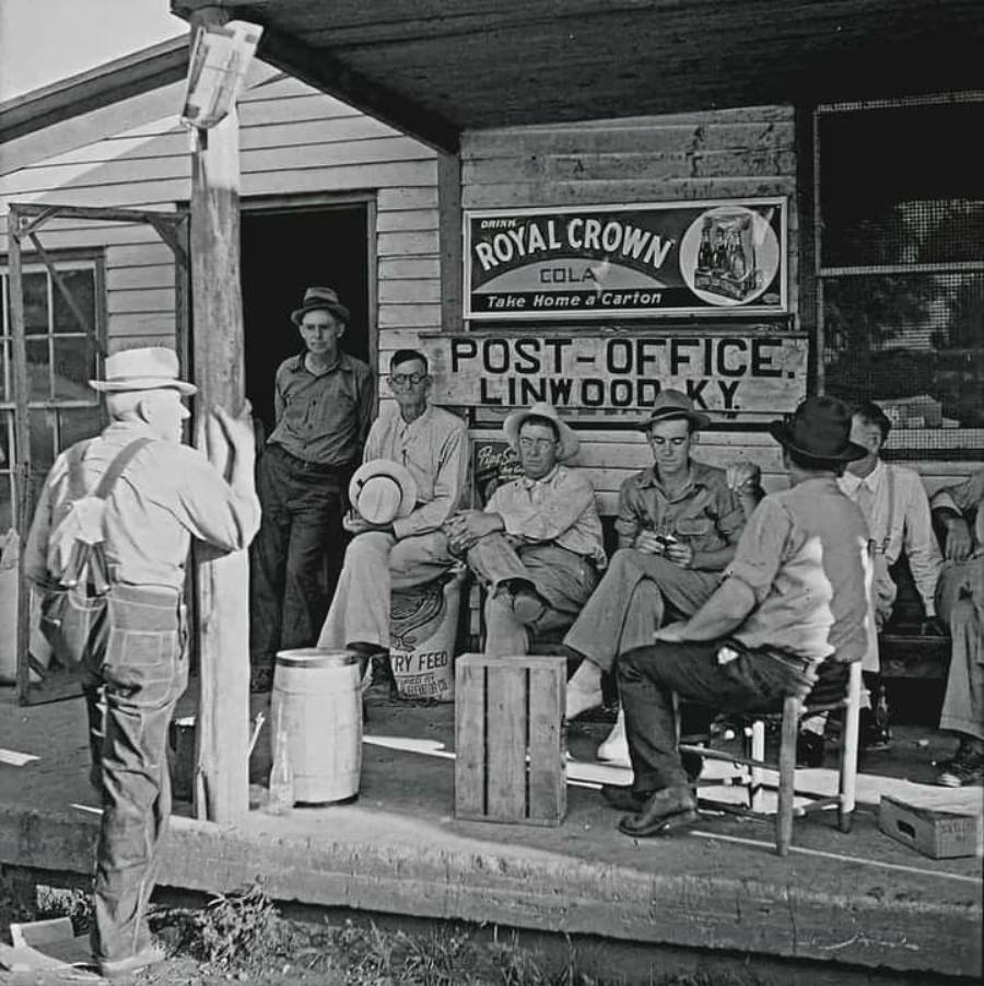 A group of men sit and stand on the porch of a rural post office and store in Linwood, Kentucky, with vintage signs visible, including one for Royal Crown Cola.