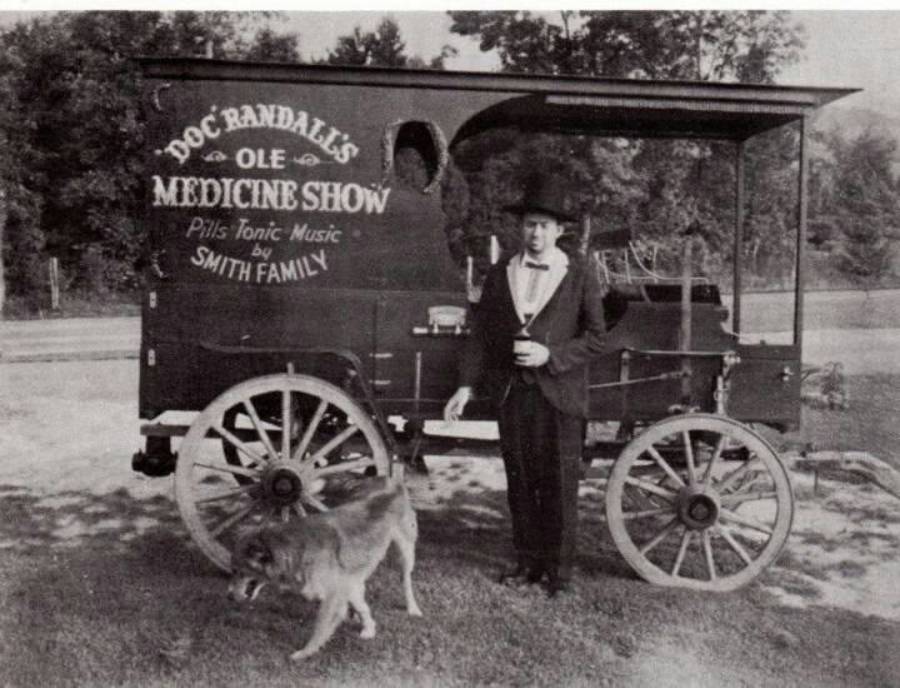 A man in vintage attire stands by a wagon labeled "Doc Randall’s Ole Medicine Show" with a dog nearby. The wagon has wooden wheels and is parked on grass with trees in the background.