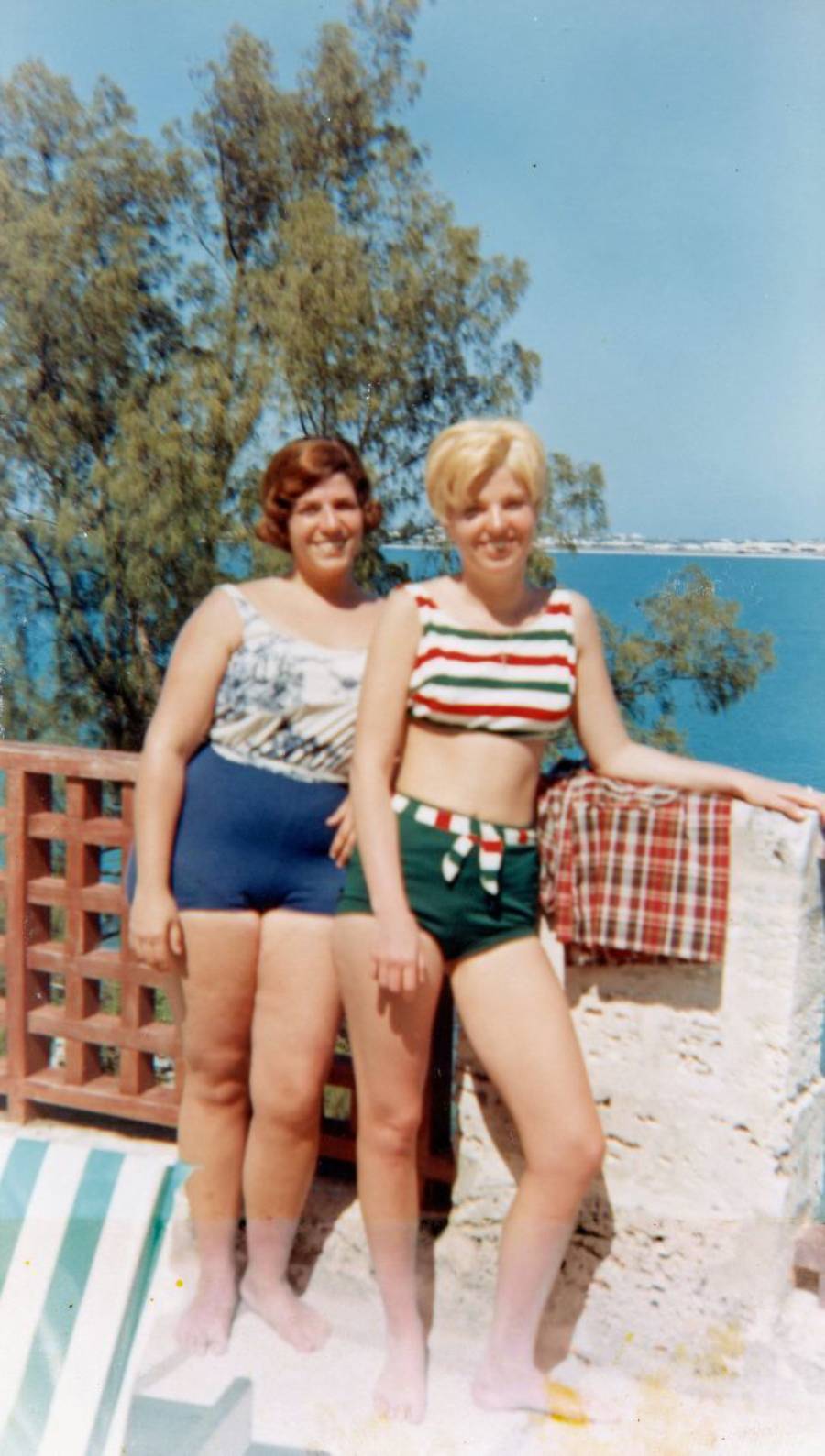 Two women in vintage swimsuits stand on a sunny patio by the sea, with trees and a blue sky in the background. One leans against a wall with a towel draped over it.