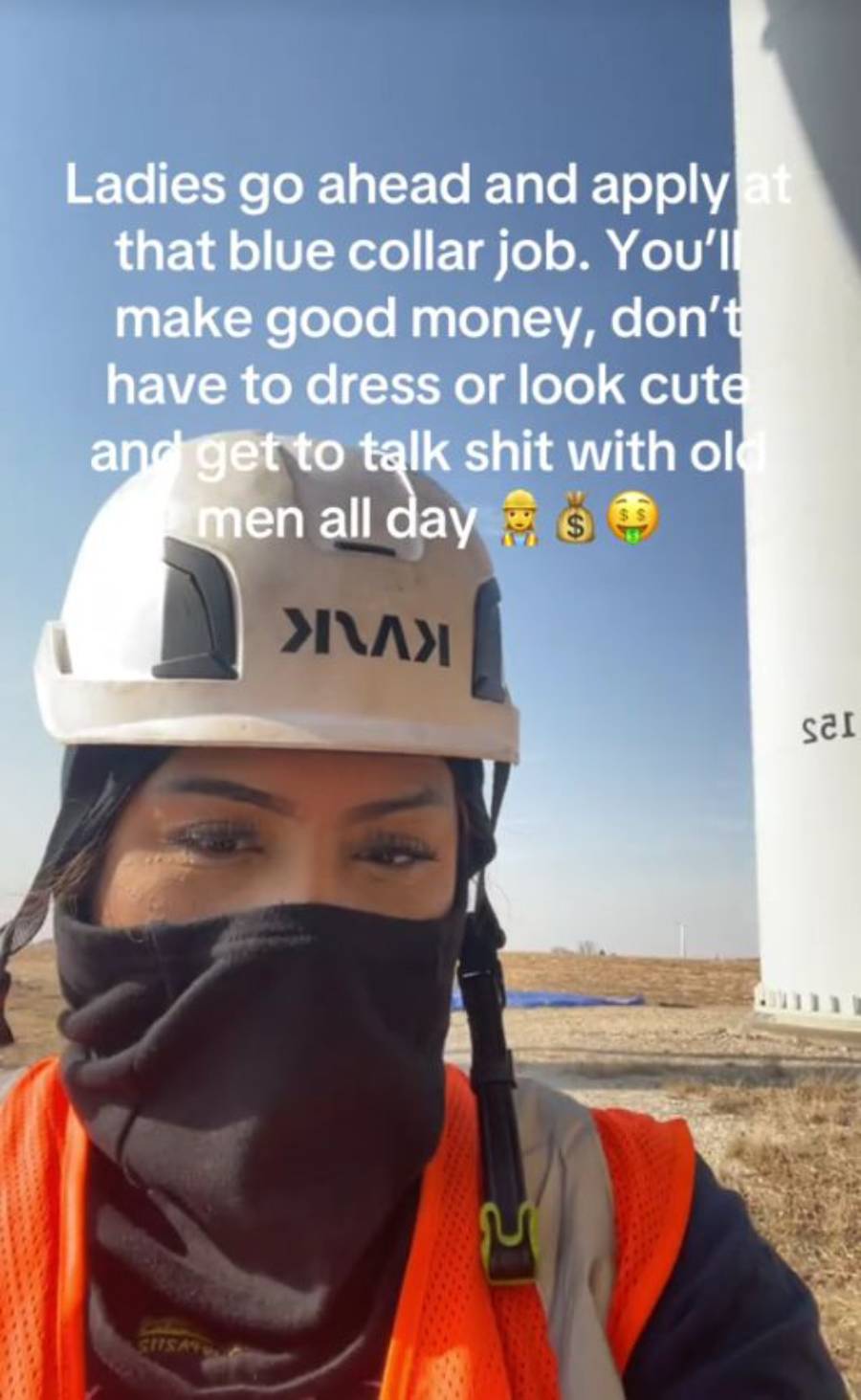 A woman in a hard hat, face covering, and safety vest takes a selfie at a worksite with a wind turbine. Text encourages women to apply for blue collar jobs for good pay and relaxed dress.