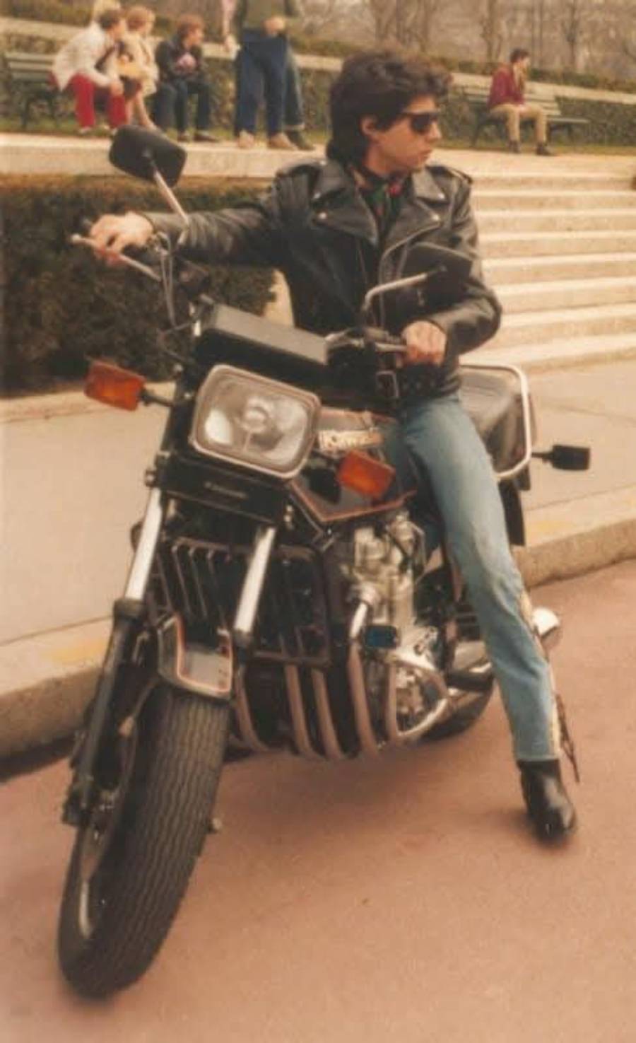 A person wearing sunglasses, a black leather jacket, and jeans sits on a motorcycle parked near some steps outdoors, with people sitting on benches in the background.