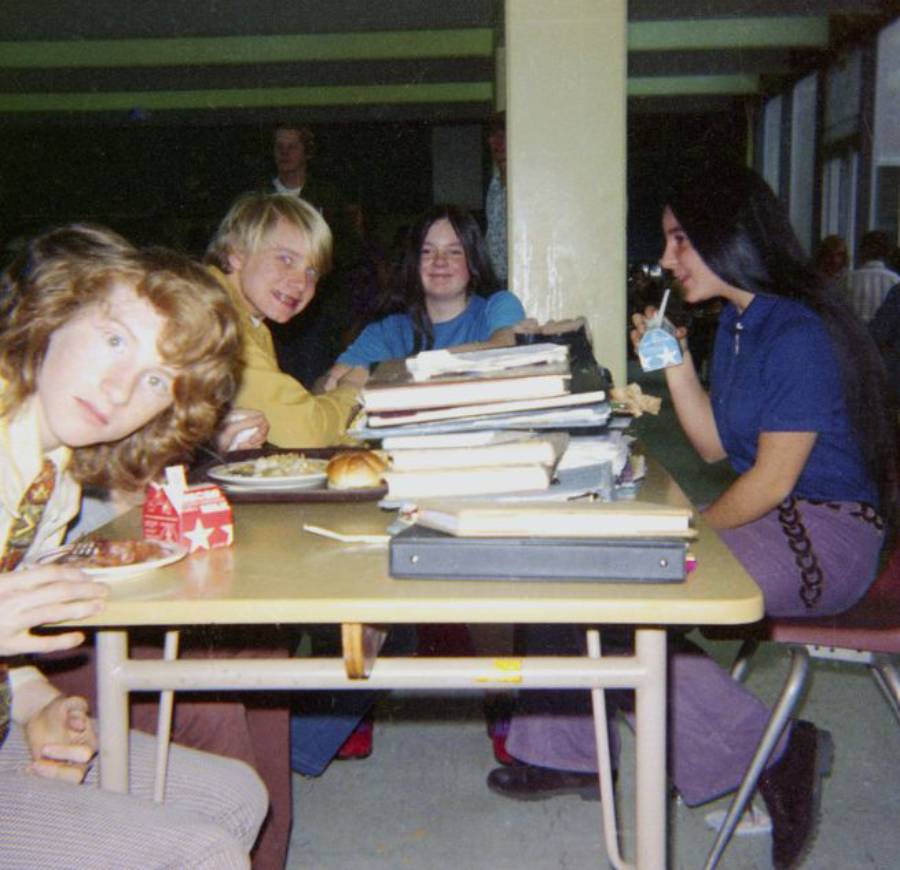 Four teenagers sit at a school lunch table with trays and milk cartons. One looks at the camera, while others are talking and eating. Books and folders are stacked high in the center of the table.