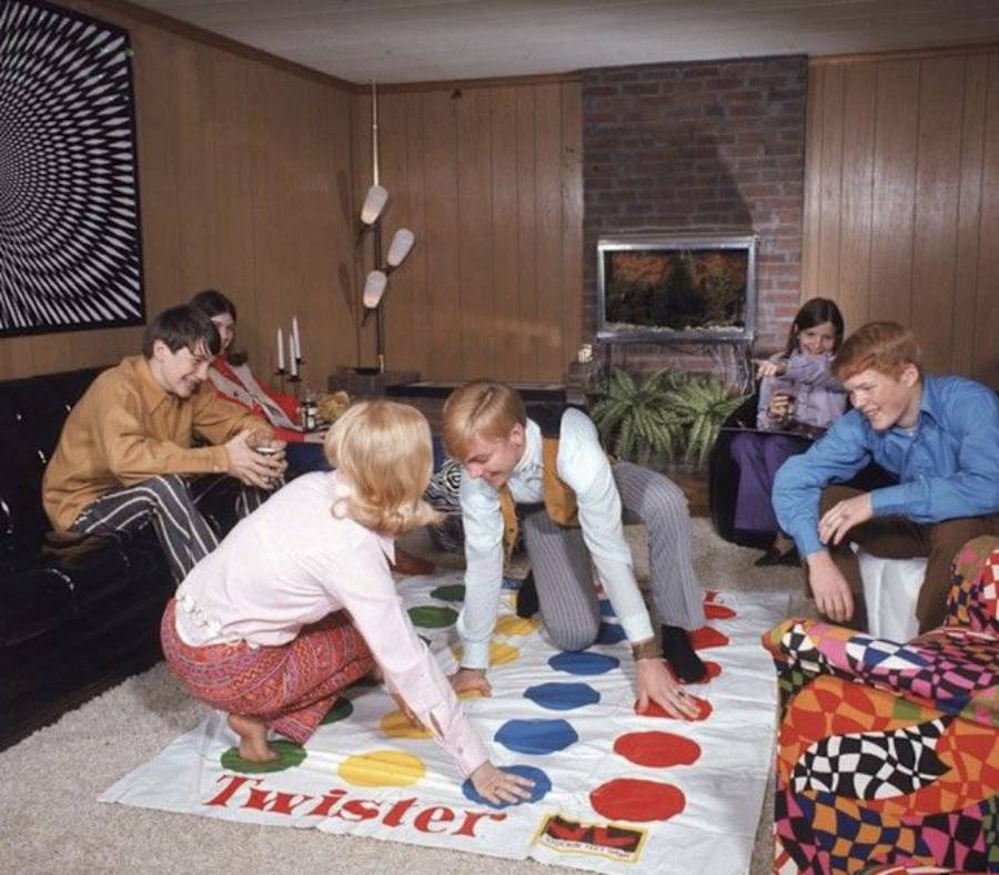 Five people in retro clothing play Twister in a wood-paneled living room. Two play the game while others watch, laughing. The room has a fireplace, plants, and bold-patterned décor.