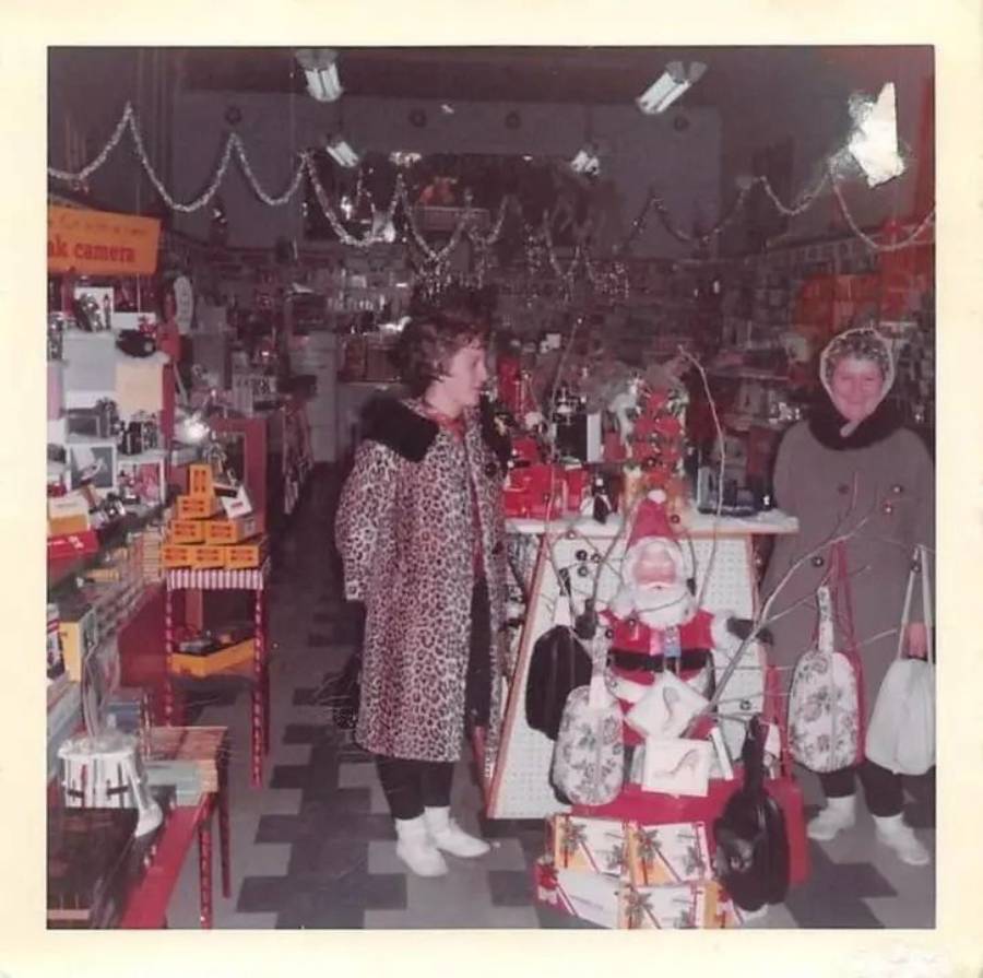 Two women in winter coats stand in a festive, cluttered store filled with products and Christmas decorations, including a Santa Claus figure on display. Shelves are stocked, and garlands hang from the ceiling.