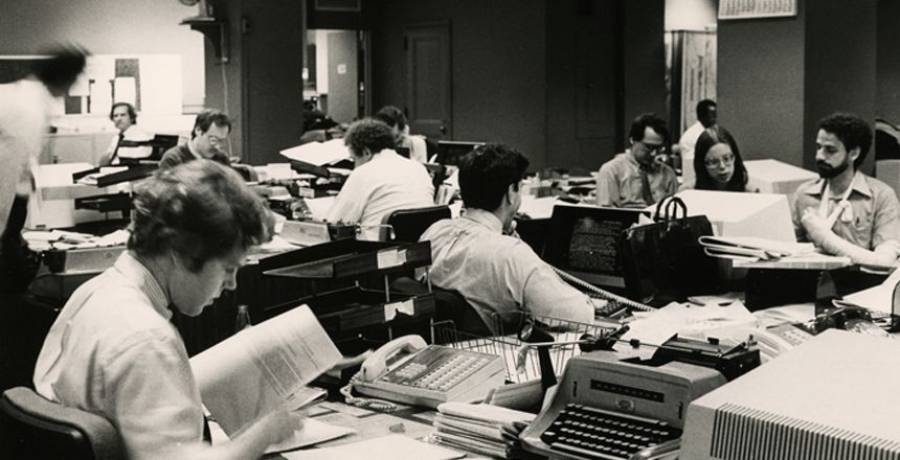 Black and white photo of a busy newsroom, with people working at desks covered in papers, typewriters, and office supplies. The scene captures a bustling office environment, likely from the mid to late 20th century.