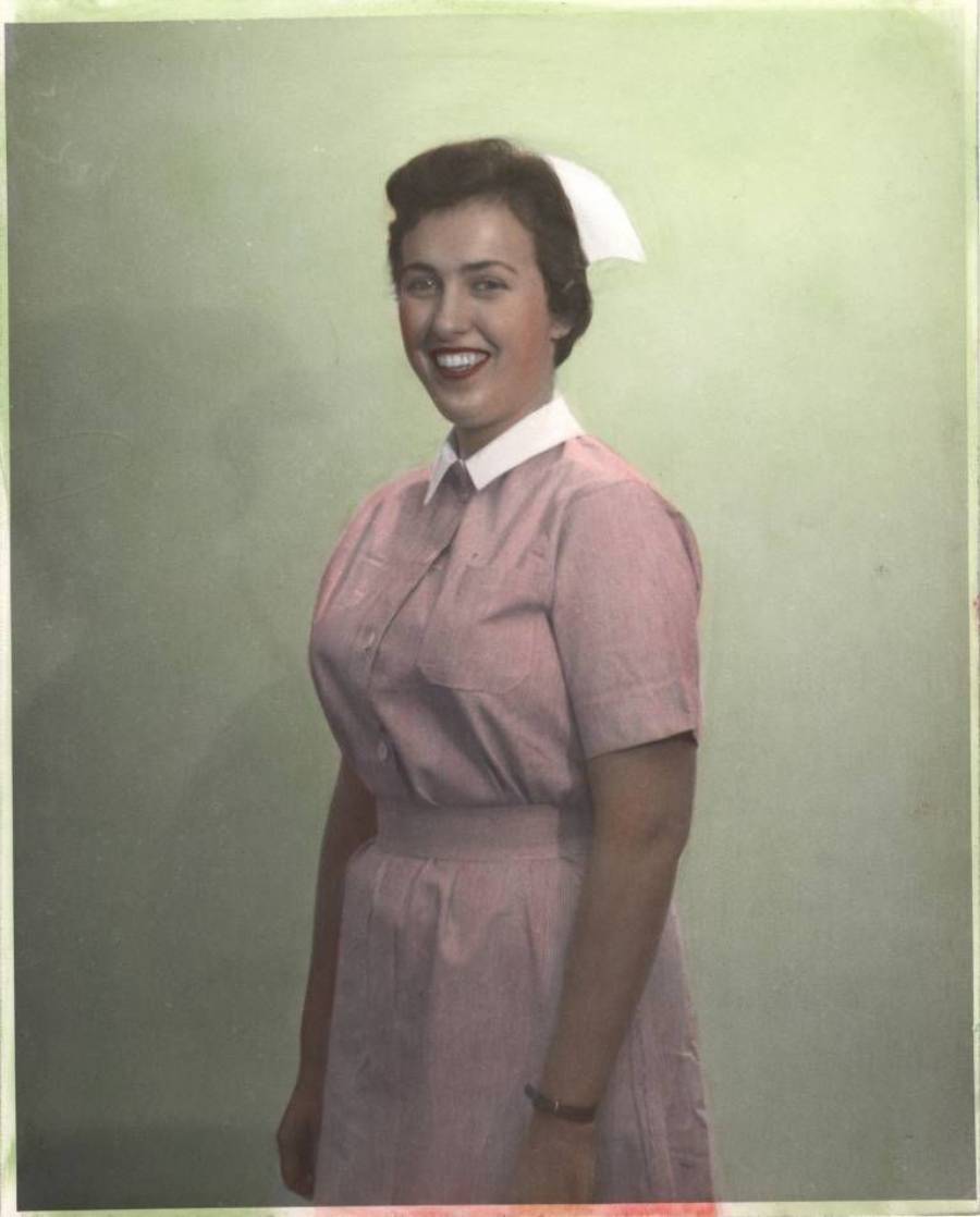 A woman in a pink nurse’s uniform with a white collar and cap stands smiling against a plain, light green background.