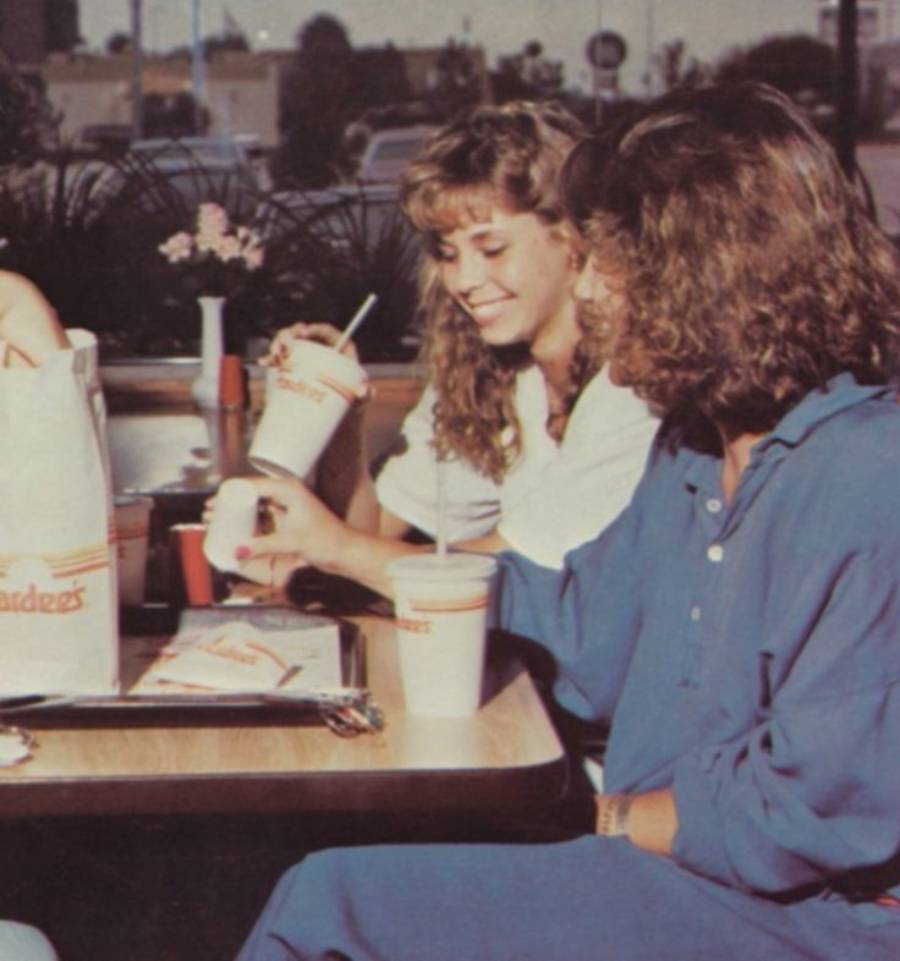 Two women sit at a restaurant table, smiling and enjoying drinks with straws. Food in branded Hardee’s packaging is on the table. It appears to be daytime, and there is a window with plants behind them.