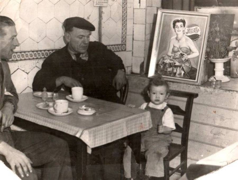 Two men sit at a café table with coffee and pastries. A young child sits nearby. On the wall, there is a framed Coca-Cola poster featuring a woman holding a bottle. The setting looks vintage and casual.