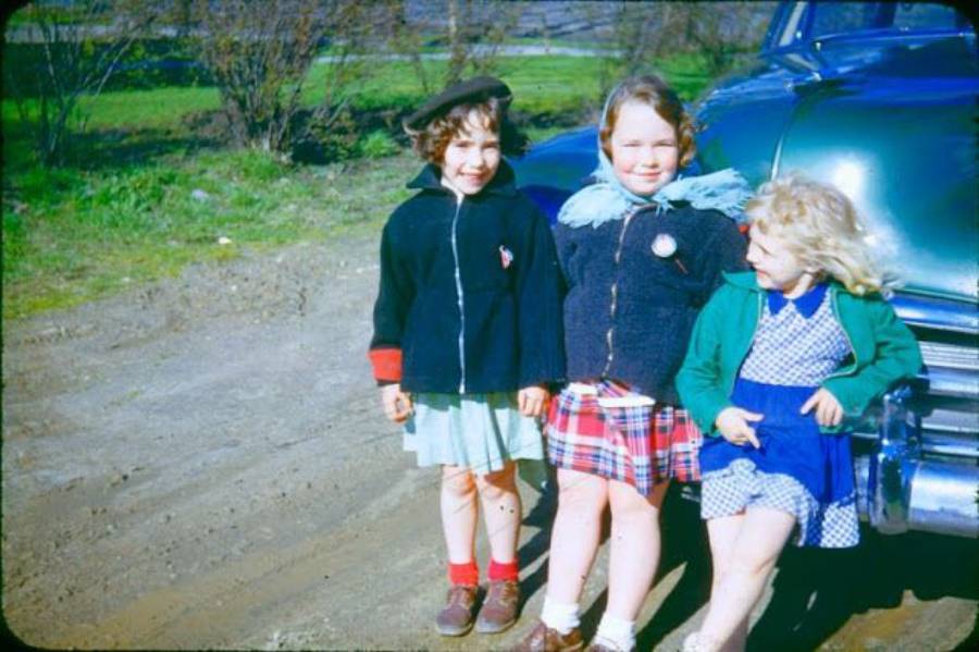 Three young girls stand smiling next to a vintage green car on a dirt road, dressed in colorful jackets and skirts, with trees and grass in the background on a sunny day.