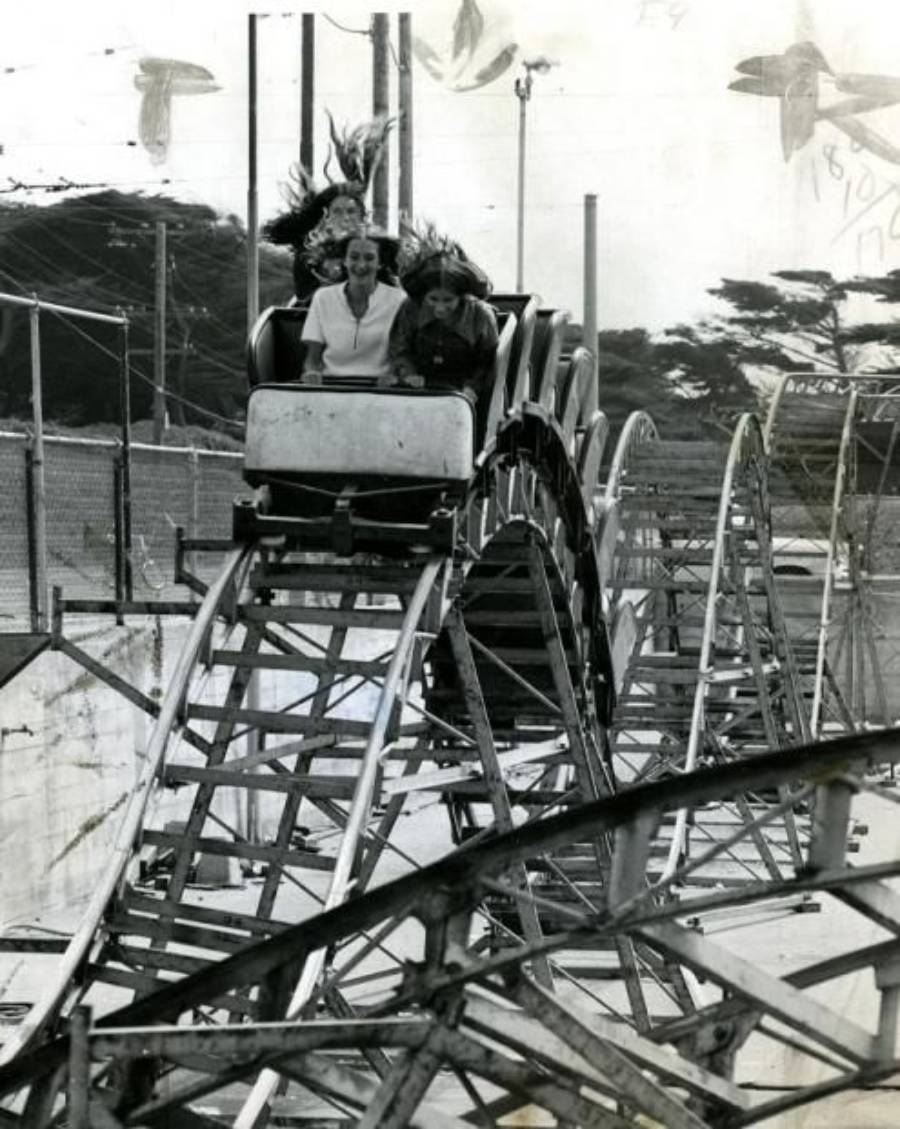 Two people ride a small, vintage roller coaster; the woman in front smiles as her hair blows back. The track features several dips and curves, with fencing and trees in the background.