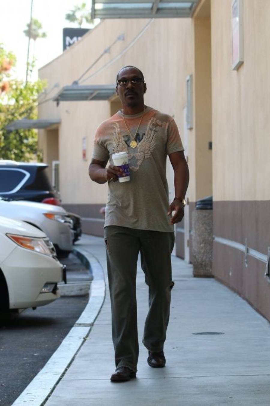A man wearing sunglasses, a t-shirt, green pants, and brown shoes walks outside on a sidewalk holding two drinks, with parked cars and a beige building in the background.