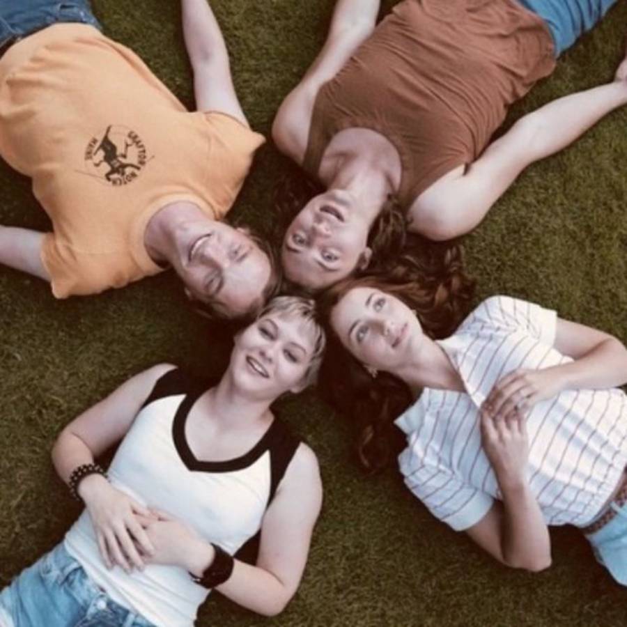Four people lie on their backs in a circle on grass, heads close together, smiling and looking up at the camera.
