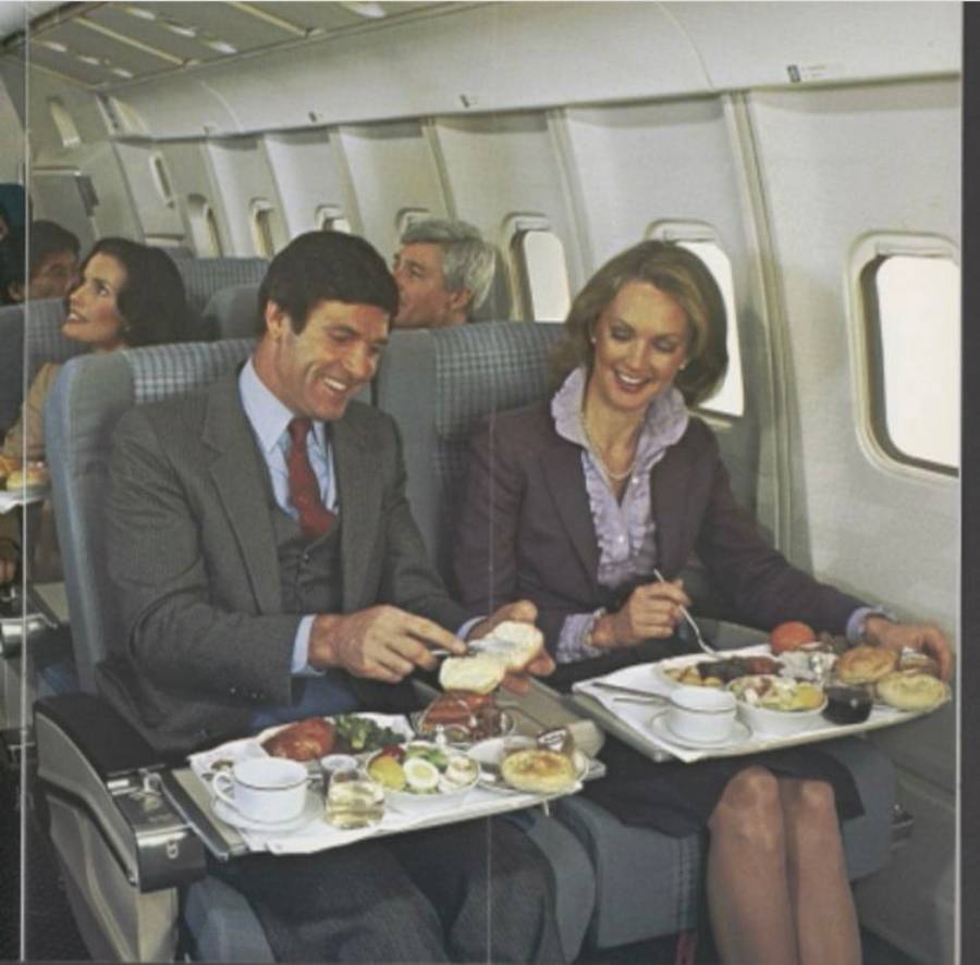 A man and woman in business attire smile while enjoying a meal on an airplane. Their trays hold various dishes. Other passengers are visible in nearby rows.