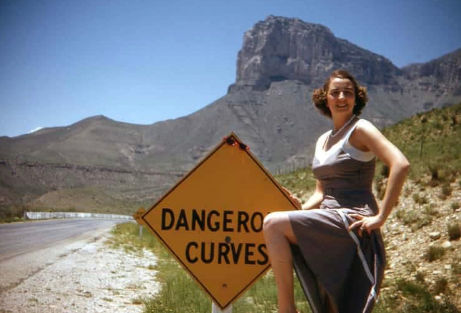 A woman in a dress poses with her leg up on a yellow “Dangerous Curves” road sign. Mountains and a clear blue sky are visible in the background.