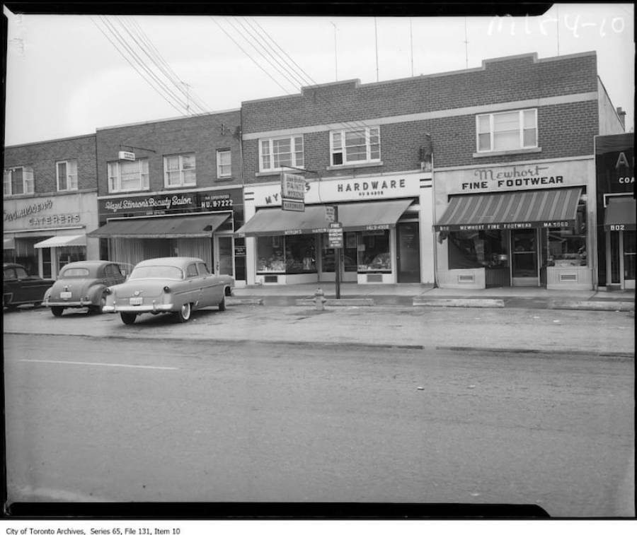 Black and white photo of a 1950s city street with parked cars in front of storefronts, including a beauty salon, hardware store, and shoe store in a brick building. Power lines are visible overhead.
