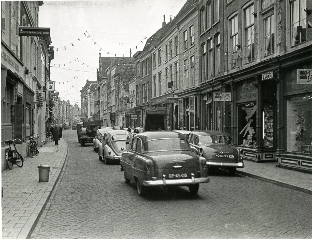 Black and white photo of a European street in the mid-20th century, with vintage cars parked and driving, bicycles on the sidewalk, shops with awnings, and string lights overhead. People walk along the sidewalks.