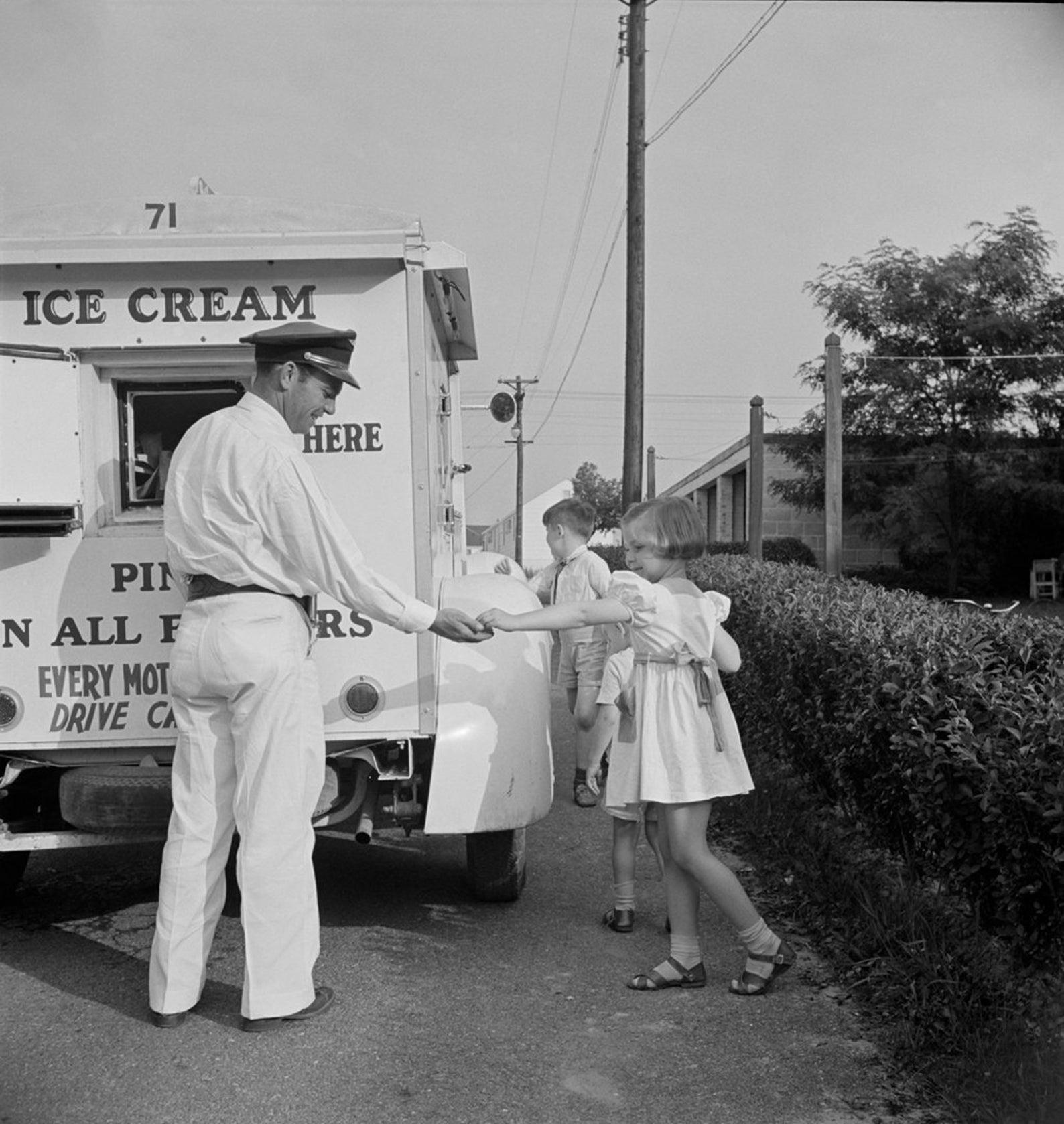 A man in a uniform serves ice cream from a truck to two children on a sidewalk. The girl in front is receiving an ice cream cone while the boy behind waits his turn. Shrubs and utility poles line the street.