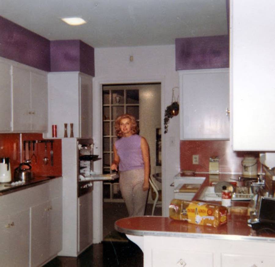 A woman with blonde hair stands in a retro kitchen with white cabinets, red countertops, and purple accents. Various kitchen items and a box of Glad Wrap are visible on the counters.