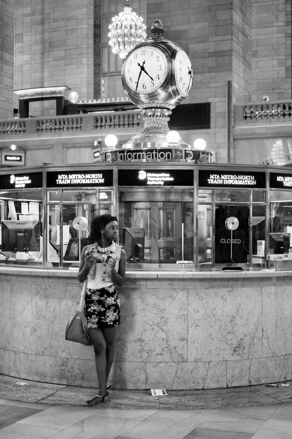 A woman in a floral skirt stands near the information booth under a large clock at Grand Central Terminal, holding a drink and looking to her left. The booth displays signs for MTA Metro-North train information.