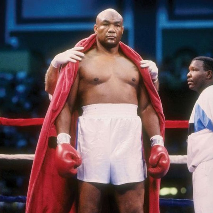 A boxer in white shorts and red gloves stands in a boxing ring, with a red robe being placed over his shoulders. Another man stands nearby, partially visible, wearing a white and blue jacket.
