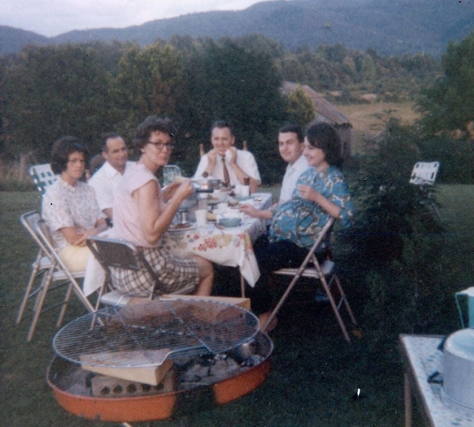Six adults sit around a table outdoors, enjoying a meal together. Trees and mountains are visible in the background, and a charcoal grill is in the foreground. Everyone appears to be smiling or talking.