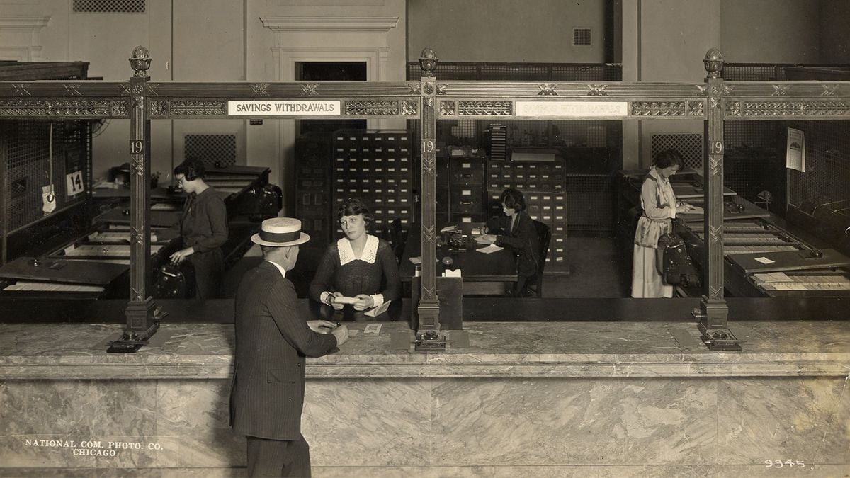 A vintage bank scene shows a man at the counter speaking to a female teller. Other women work behind barred windows, handling documents. Signs read “Savings Withdrawals.” The setting is formal, with marble counters and wood paneling.