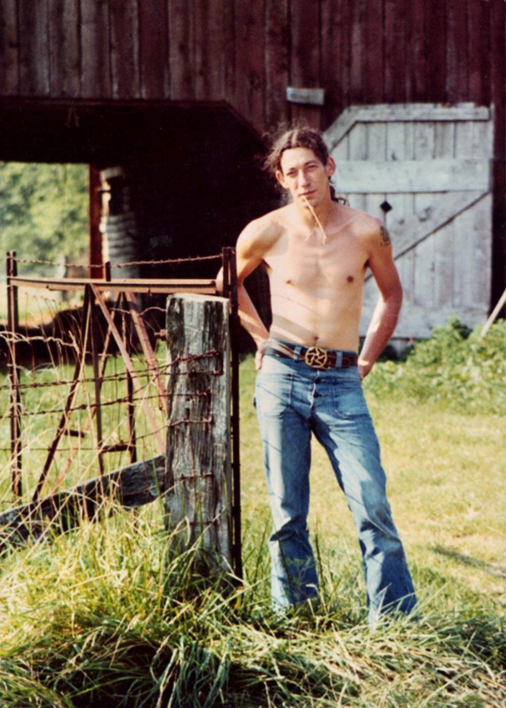 A shirtless young man in jeans stands outdoors by a wire fence, hands in pockets, with a rustic wooden barn in the background on a sunny day.