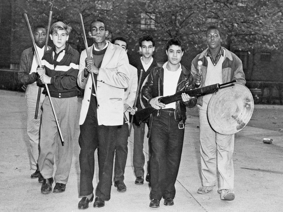 A group of seven young men walk together outdoors, carrying sticks, a bat, and a trash can lid. They appear determined and alert, moving as a unit, with trees and a building visible in the background.