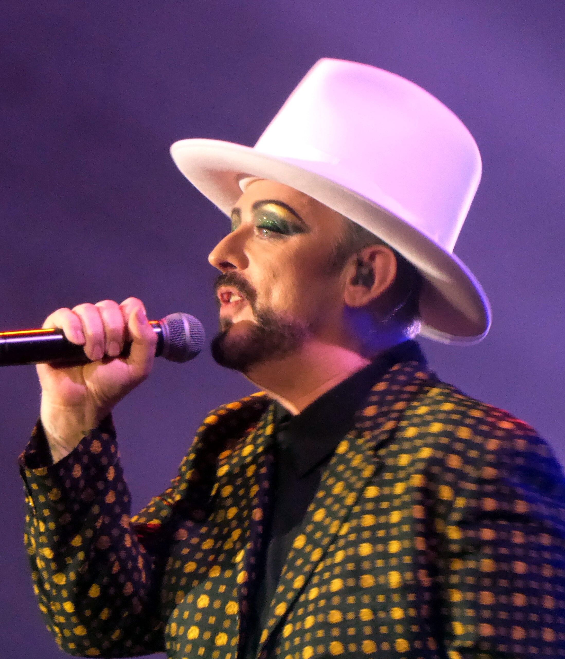 A person wearing a large white hat, patterned jacket, and dramatic makeup sings into a microphone on stage under colorful lighting.