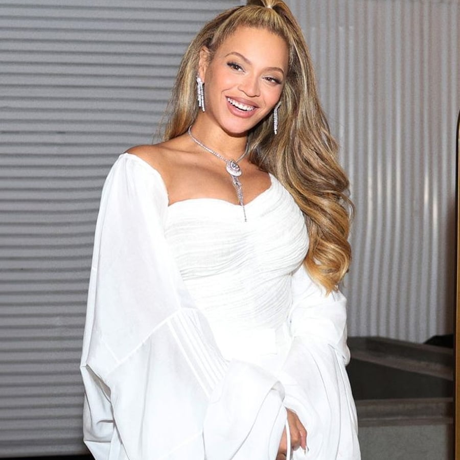 A woman with long, wavy blonde hair in a high ponytail smiles, wearing a white off-the-shoulder dress and silver jewelry, standing in front of a ribbed white background.