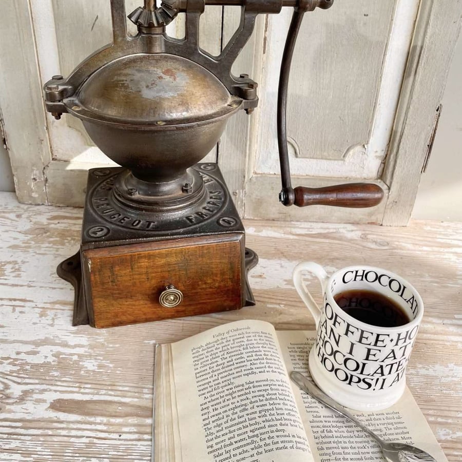 Vintage coffee grinder, a white mug with coffee, and an open book rest on a light surface against a rustic wooden background. A spoon lies atop the open book near the mug.