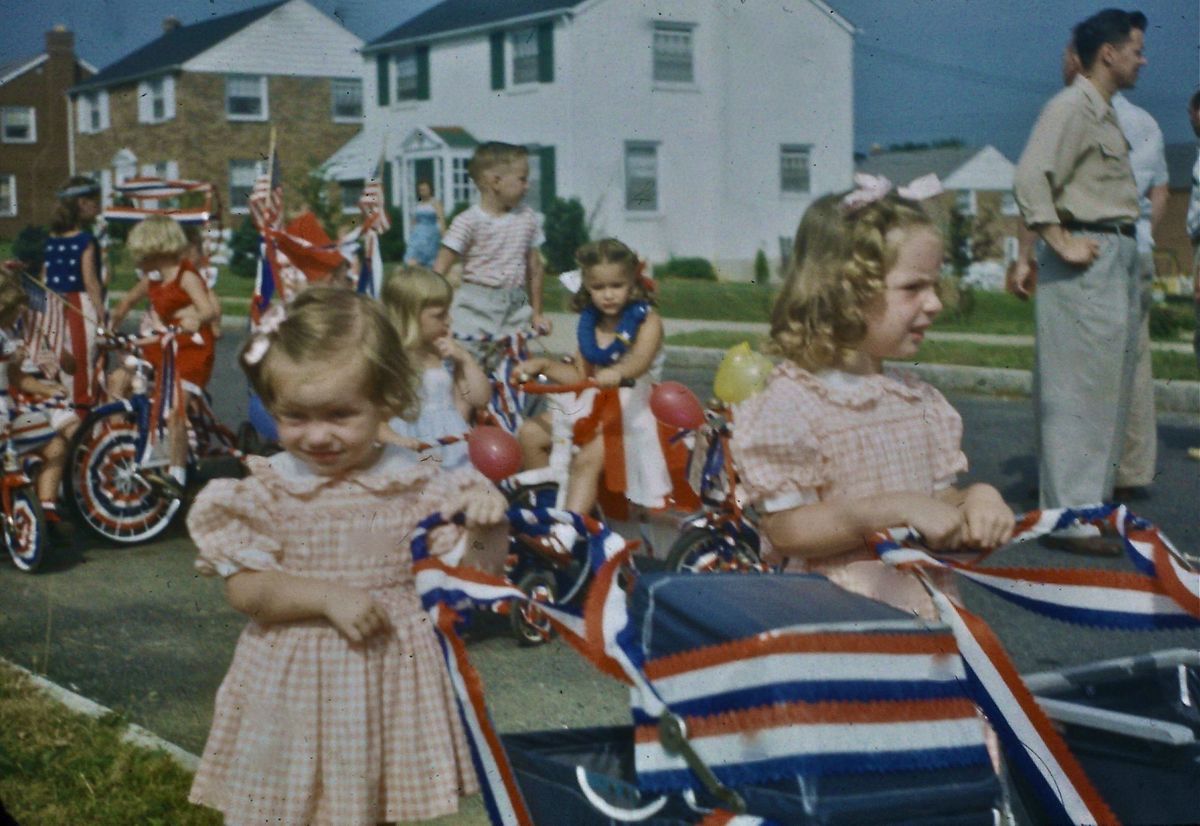 Young children in matching pink dresses hold red, white, and blue streamers at a parade. Decorated bikes and houses line the background, with adults and other children nearby.
