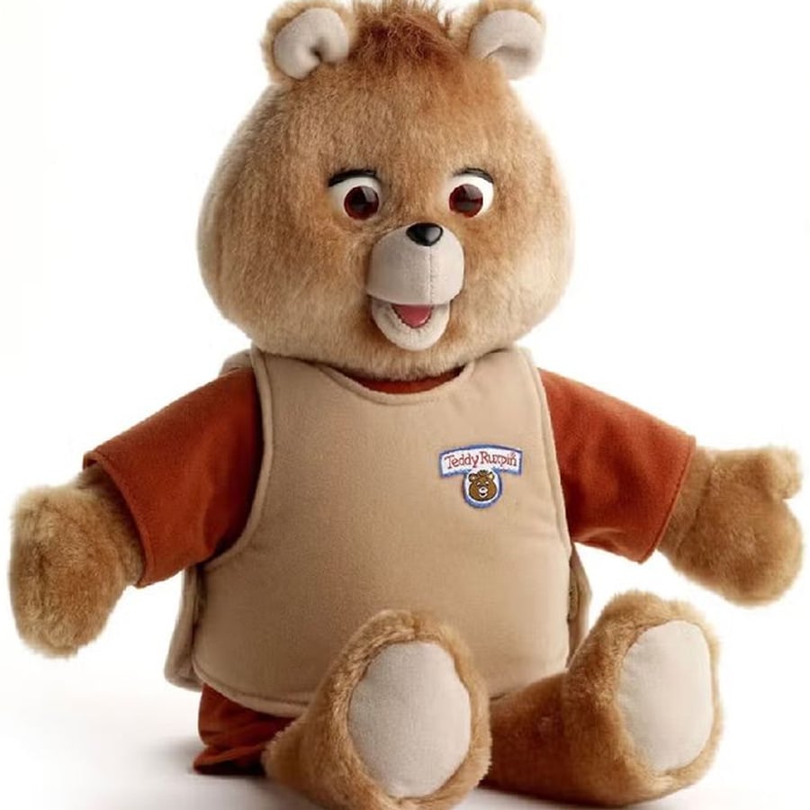 A brown teddy bear toy with a tan vest labeled "Teddy Ruxpin," brown eyes, tan snout, and tan paws, sitting upright against a white background.