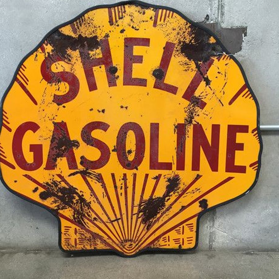 A vintage, weathered Shell Gasoline sign shaped like a yellow scallop shell, with red letters. The sign has black stains and is mounted against a concrete wall.