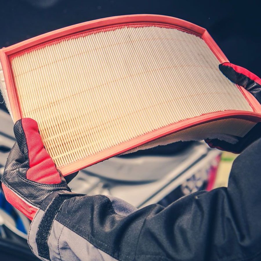 A person wearing gloves holds a clean car air filter with a red frame, inspecting it over an open engine bay.
