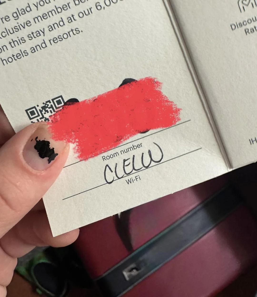 A hand holding a booklet with a red mark covering some text. Below, handwritten text reads "CLEM" under "Room number." The person's nails are chipped with black nail polish.