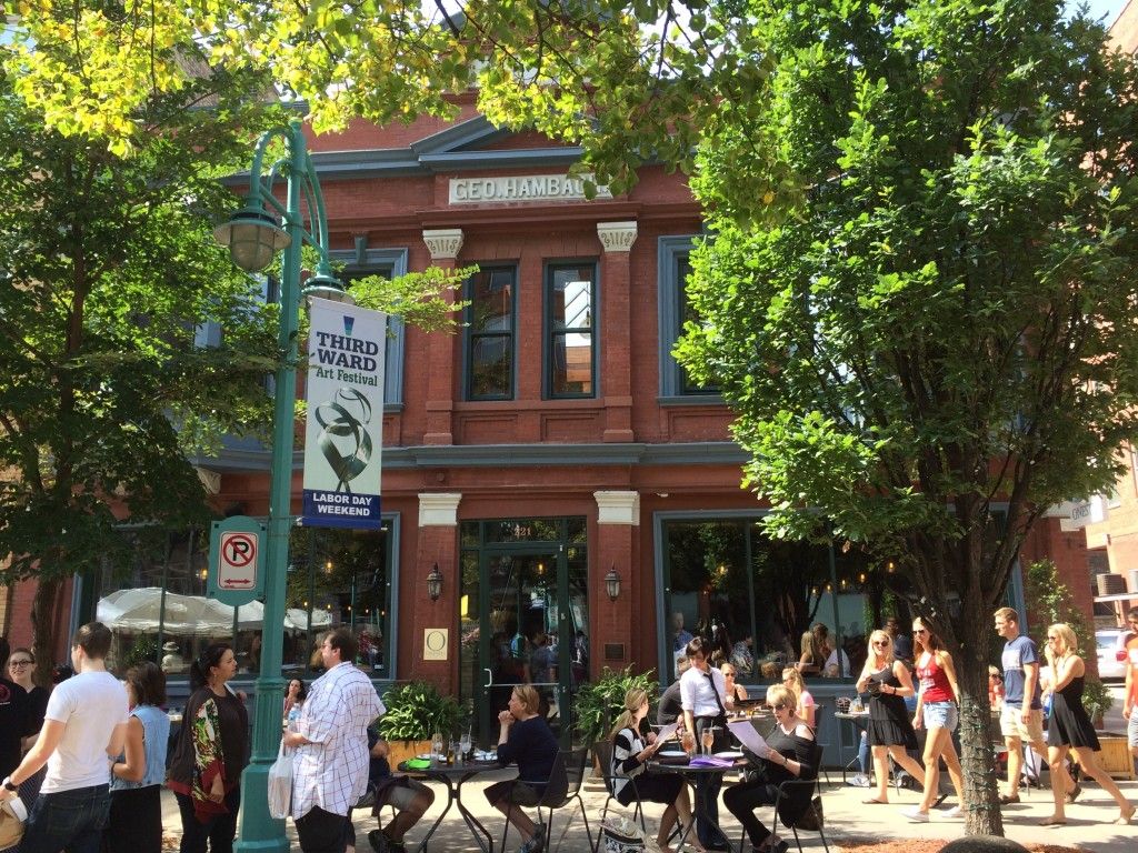 People gather outside a red-brick building with large windows, trees, and outdoor tables. A sign advertises the Third Ward Art Festival on Labor Day weekend. Sunlight filters through the trees, creating a lively street scene.