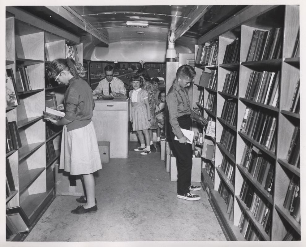 Black and white photo of children browsing books inside a bookmobile, with shelves lining both walls and a librarian standing at the back counter.