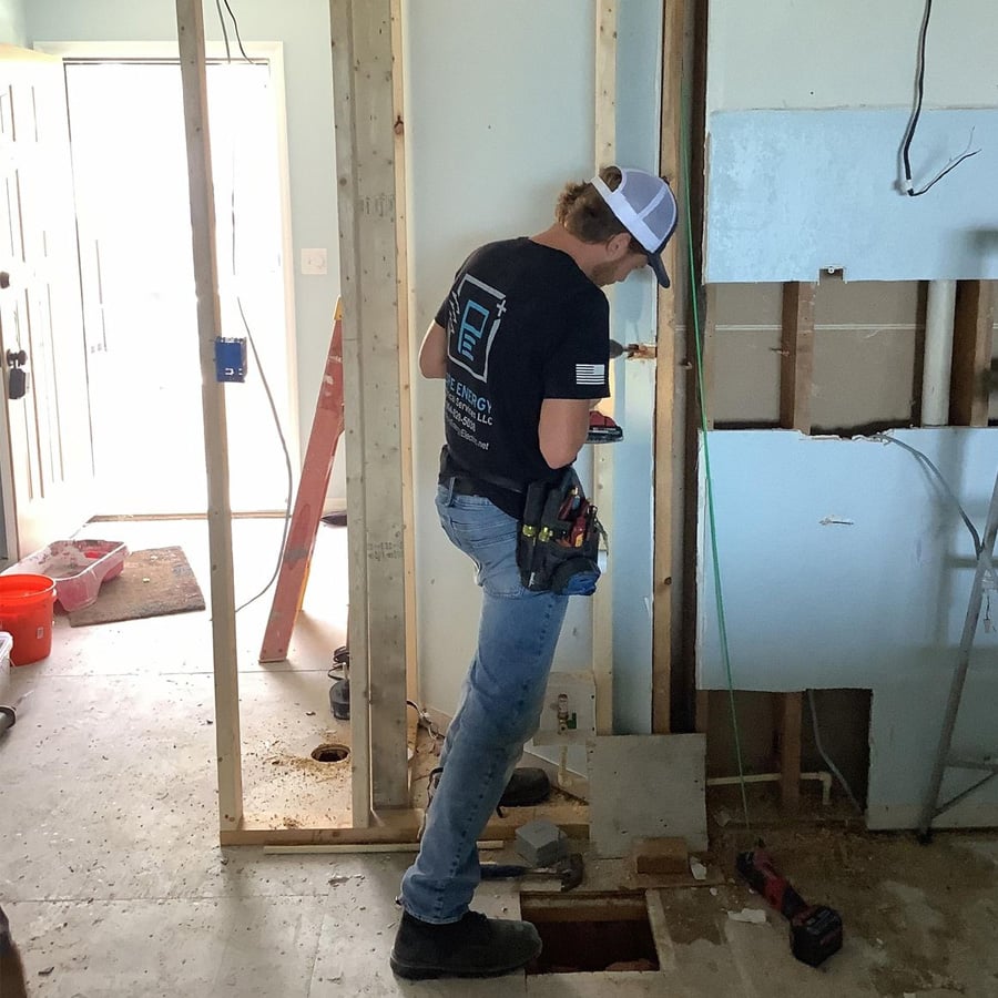 A construction worker wearing a cap and tool belt stands in a partially finished room, working with electrical wiring in wooden wall framing. Tools and equipment are scattered around the site.