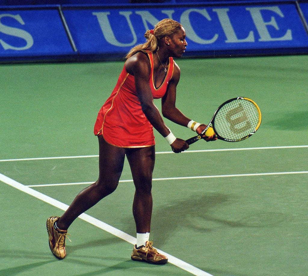 A female tennis player in a red dress prepares to hit a backhand shot on a green hard court during a match.
