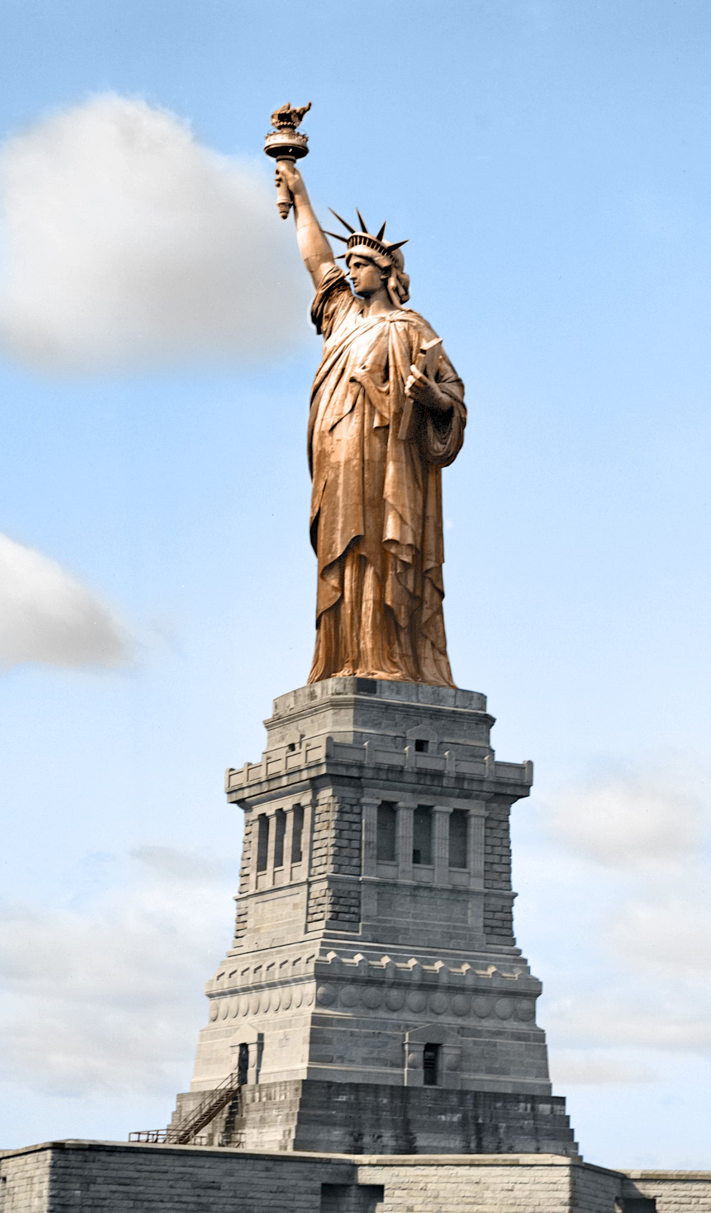 The Statue of Liberty stands on its stone pedestal against a blue sky with scattered clouds, holding a torch in her right hand and a tablet in her left.