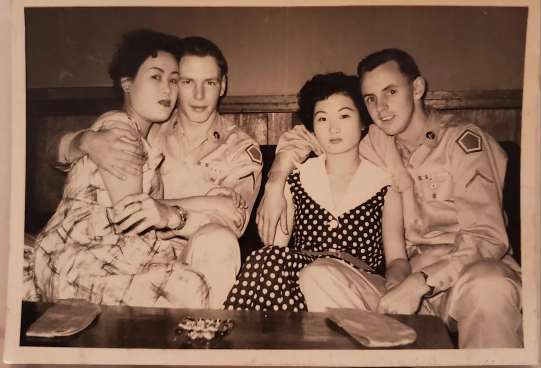 Two men in military uniforms sit closely with two women in dresses at a table, posing together and smiling for a black and white photo, likely from the 1950s.
