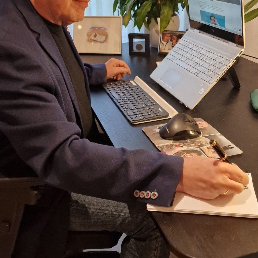 A person in a blue blazer writes in a notebook at a desk with a keyboard, mouse, laptop, framed photos, and a plant.