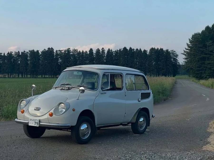 A vintage white Subaru 360 car is parked on a rural gravel road, with green fields on both sides and a forest of tall trees in the background under a clear sky.